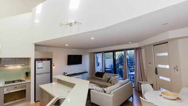 Cocina moderna con estufa a gas, refrigerador y horno empotrado. Hay un comedor con mesa y sillas, y un sofá en la sala de estar. Un televisor de pantalla plana está montado en la pared.