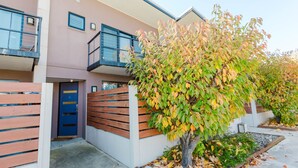Un edificio moderno de dos pisos con un balcón, una puerta de madera y un árbol con hojas otoñales.