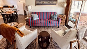Ein modernes Wohnzimmer mit einem gestreiften Sofa, zwei weißen Sesseln, einem Couchtisch und einer Küchenzeile im Hintergrund.