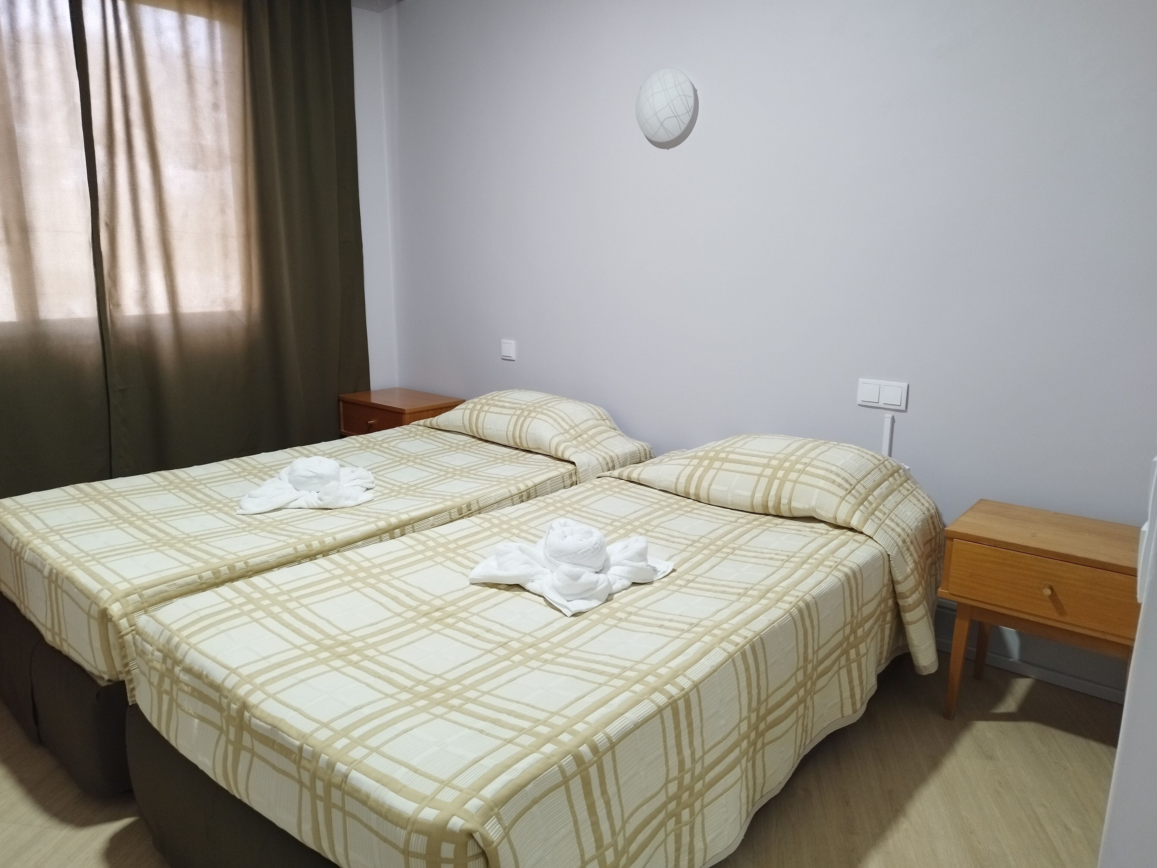 Habitación de hotel con dos camas, cada una con cubrecama a cuadros, una mesita de noche, un cajón de madera y un reloj de pared.