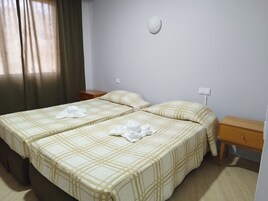 Ein Hotelzimmer mit zwei Betten, jeweils mit kariertem Überwurf, einem Nachttisch, einer Holzkiste und einer Wanduhr.