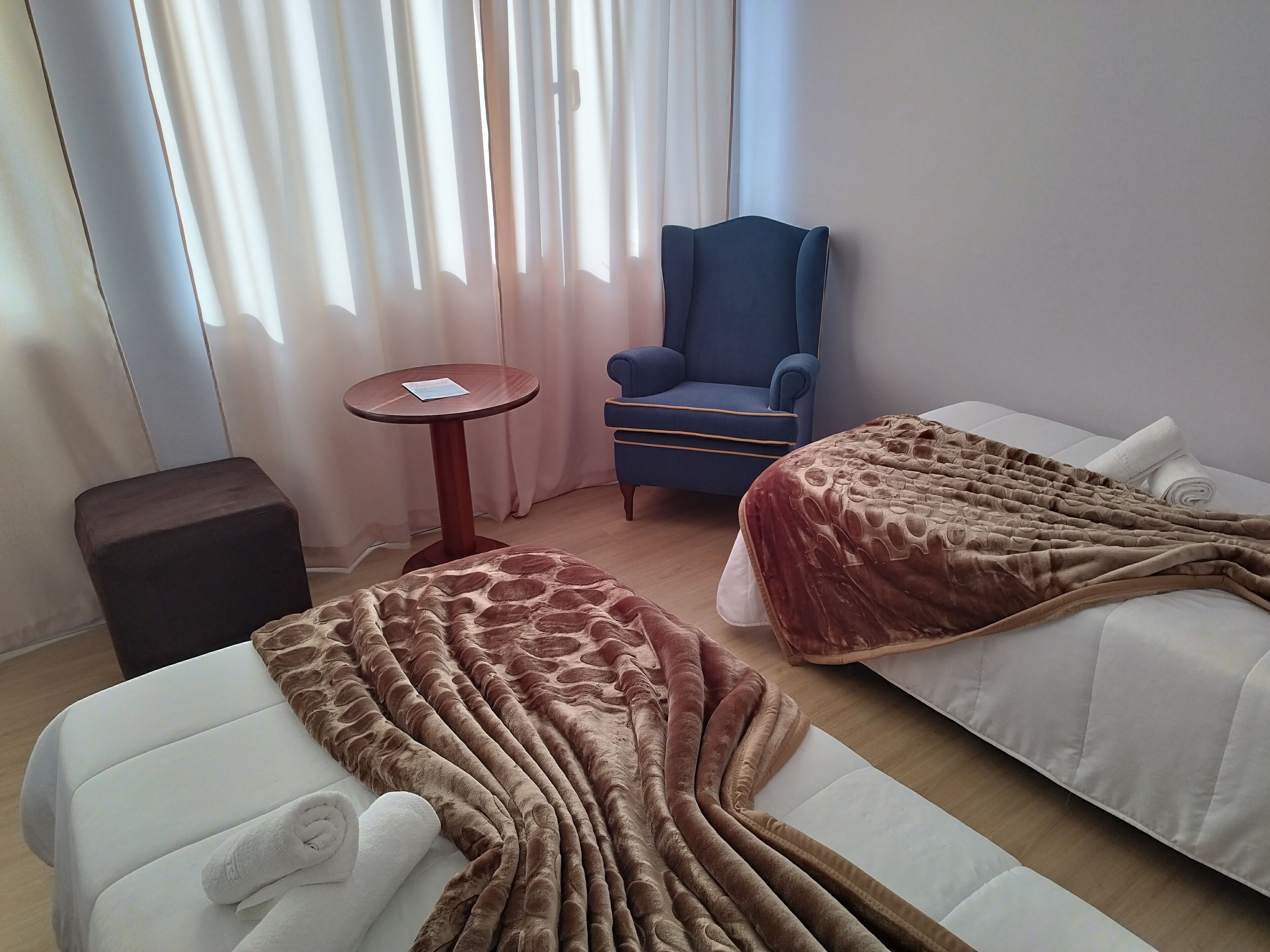Una camera d'albergo con due letti, un tavolino rotondo, una poltrona blu e un poggiapiedi scuro.