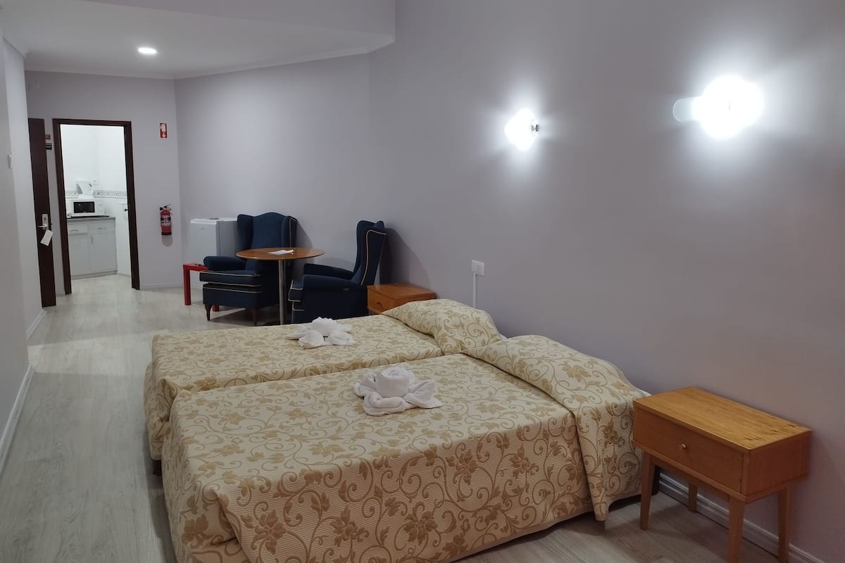 Une chambre d’hôtel avec deux lits, une petite table, une chaise, une table de chevet en bois et, au fond, une kitchenette.