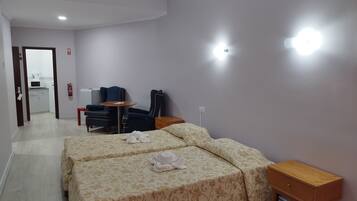 A hotel room with two beds, a small table, a chair, a wooden nightstand, and a kitchenette in the background.