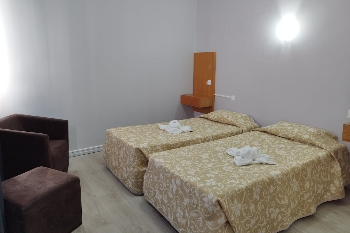 Une chambre d’hôtel avec deux lits, une table de chevet en bois, une chaise et une étagère fixée au mur.