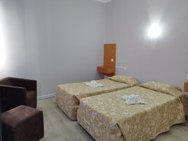 Ein Hotelzimmer mit zwei Betten, einem hölzernen Nachttisch, einem Sessel und einem an der Wand befestigten Regal.