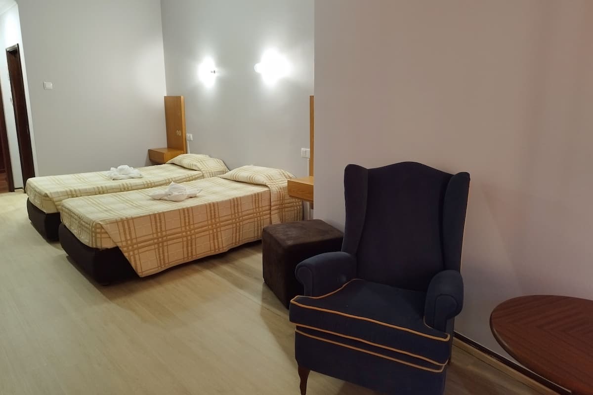 Une chambre d’hôtel avec deux lits, un fauteuil sombre, une petite table en bois et un luminaire fixé au mur.