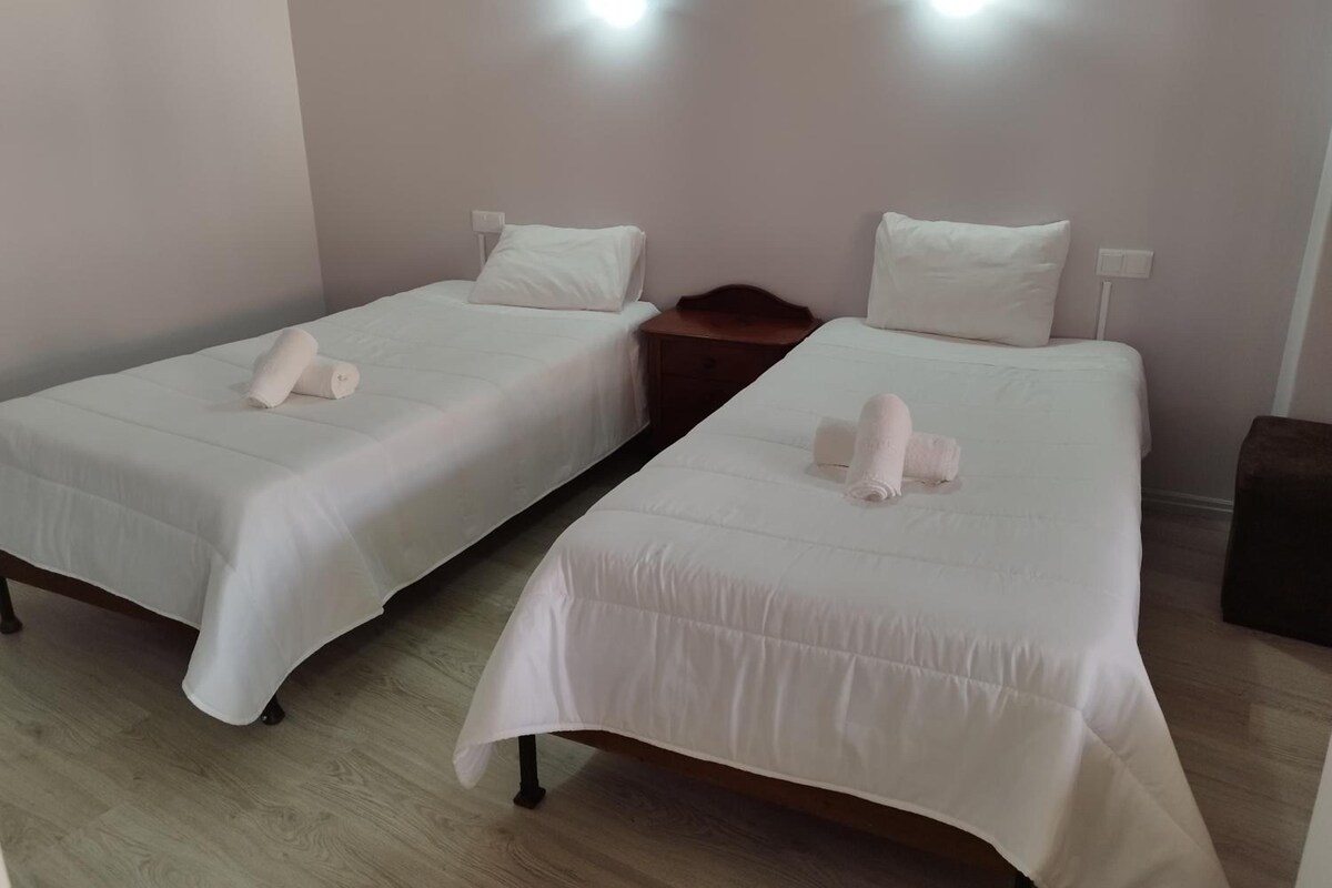 Deux lits simples avec du linge de lit et des oreillers blancs, une table de chevet en bois et un luminaire fixé au mur.