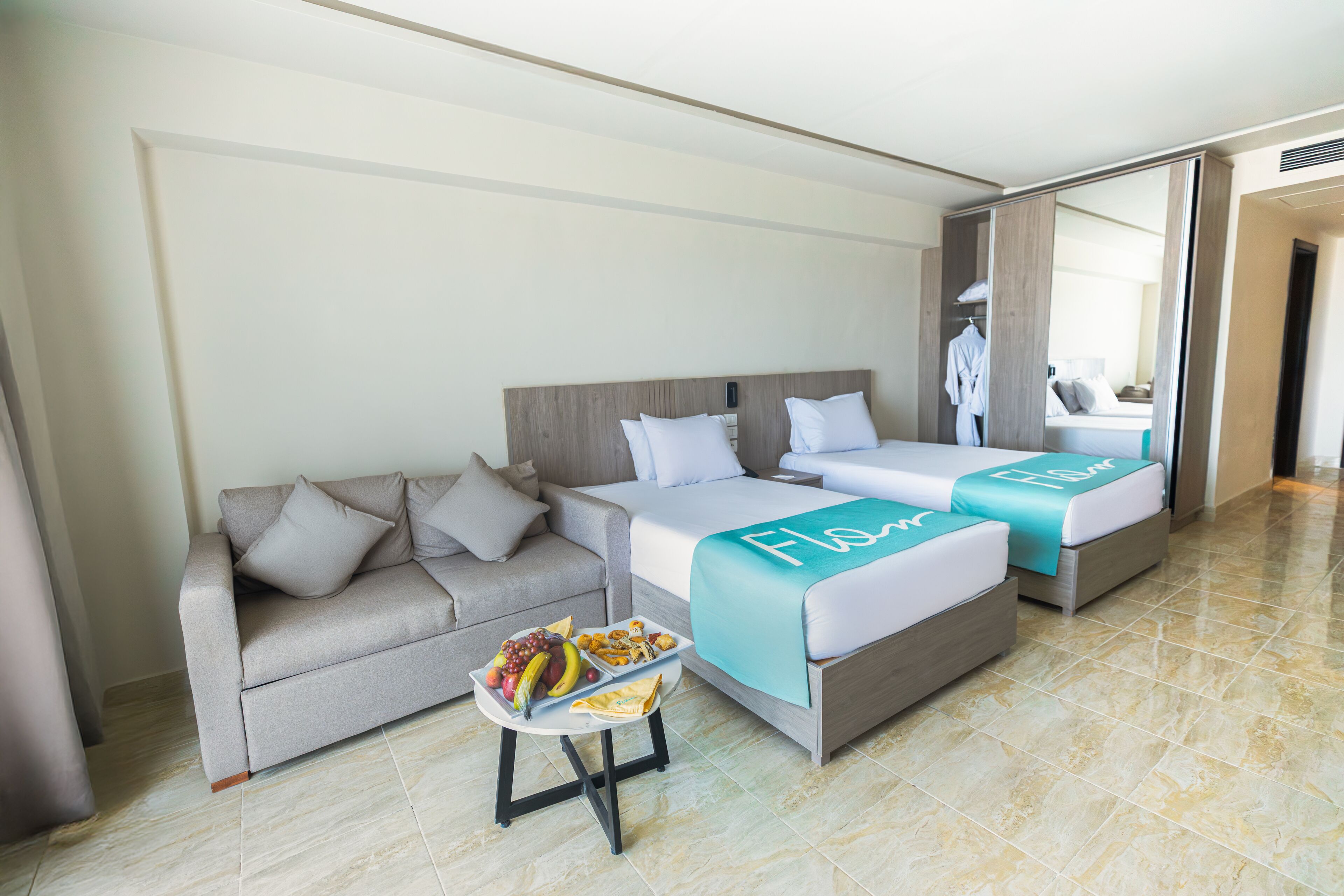 A hotel room with two beds, a sofa, a small table with fruit, and a mirror.