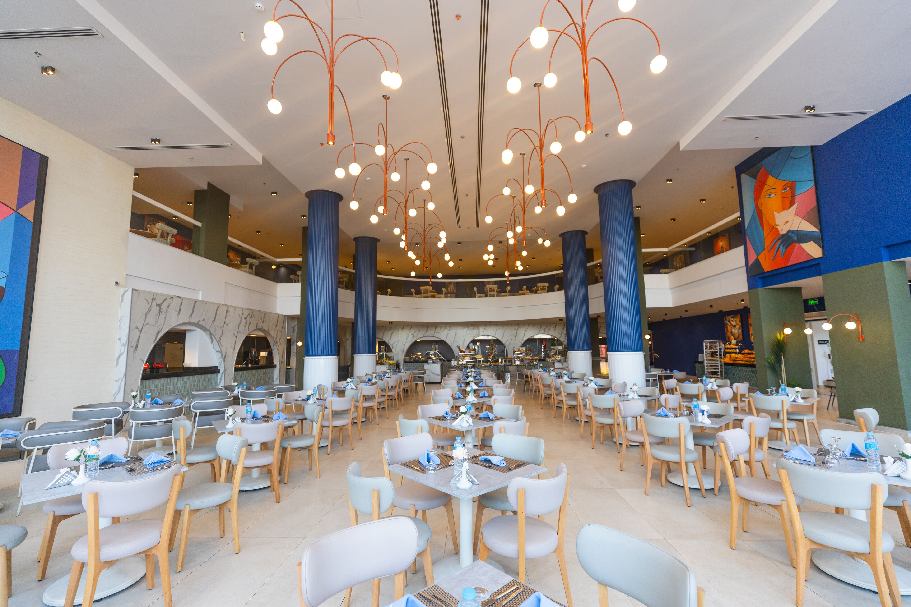 A spacious dining area with multiple tables and chairs, high ceilings with hanging lights, and a central decorative feature.