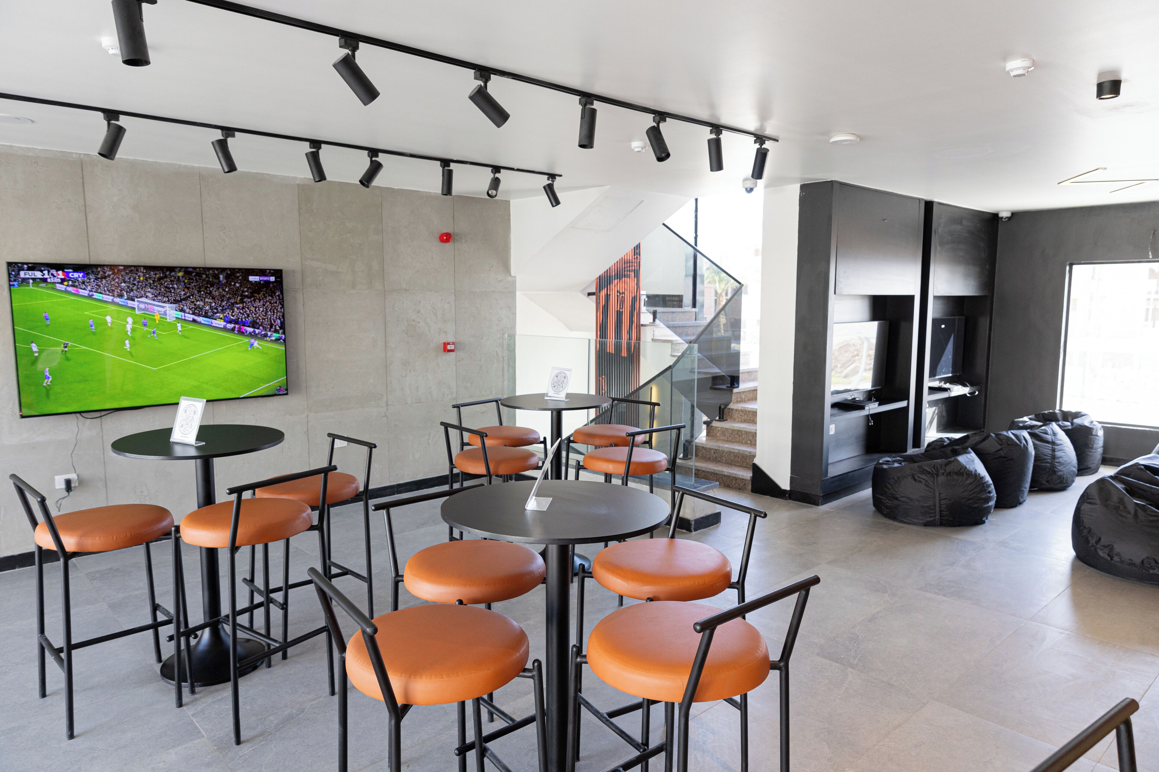 A modern lounge area with a large TV screen displaying a soccer match, round tables with orange chairs, and a staircase leading to an upper level.