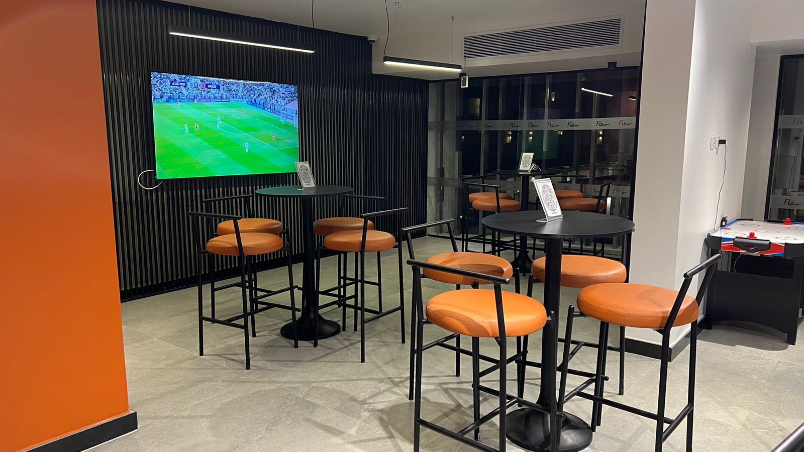 Un espace bar moderne avec des tables rondes et des chaises orange, un grand écran de télévision diffusant un événement sportif et une table de baby-foot.
