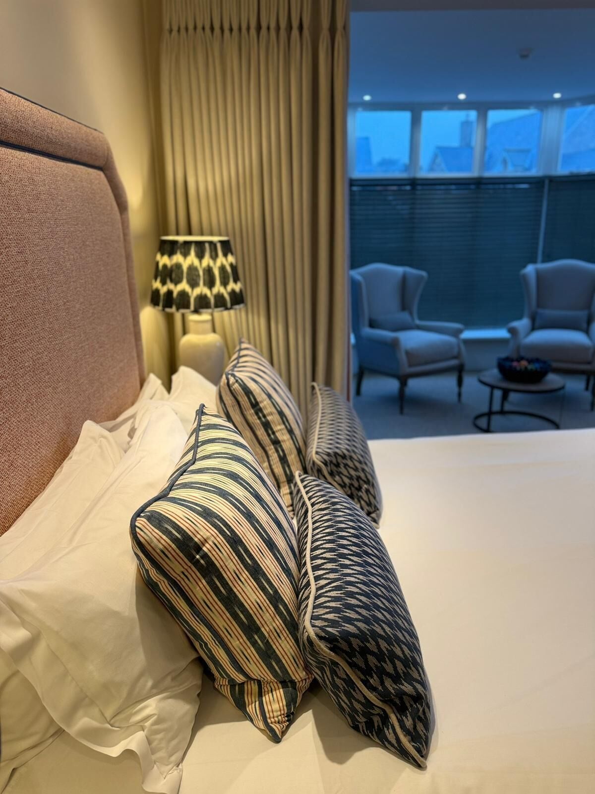 A hotel room with a large bed, striped pillows, a lamp, and a view of the city through the window.