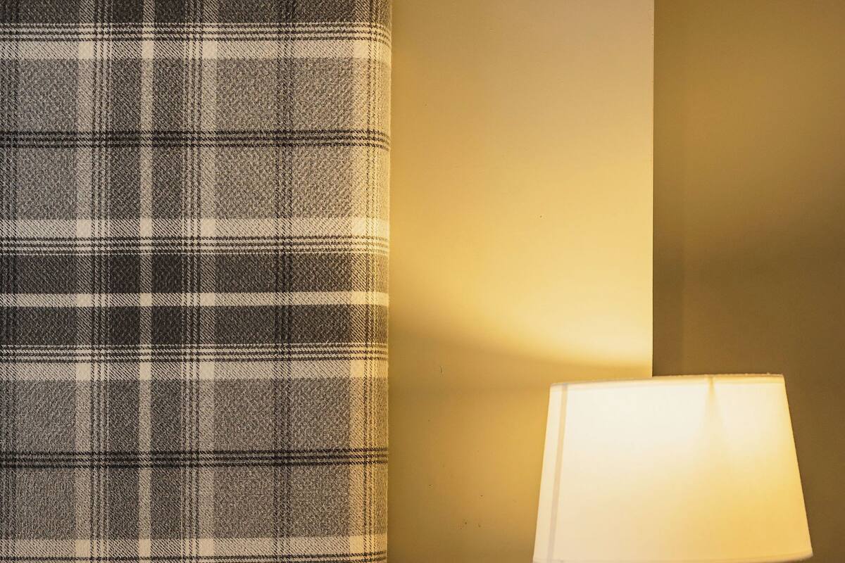 Una cama con sábanas blancas, cabecera de cuadros y una lámpara de noche sobre una mesita de madera.