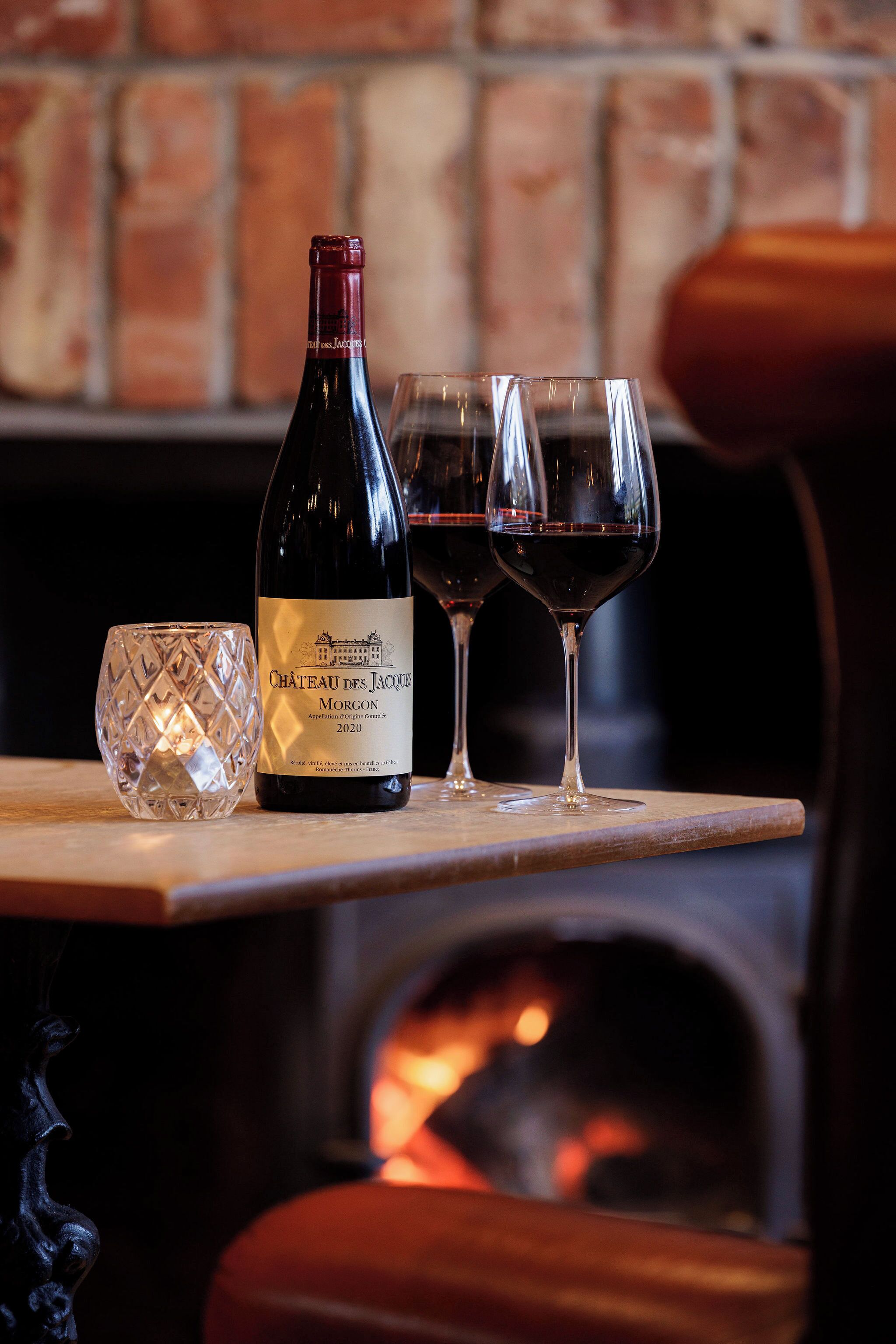 Una botella de vino Chateau des Jacques Morgon 2020, dos copas de vino y una copa decorativa sobre una superficie de madera cerca de una chimenea.