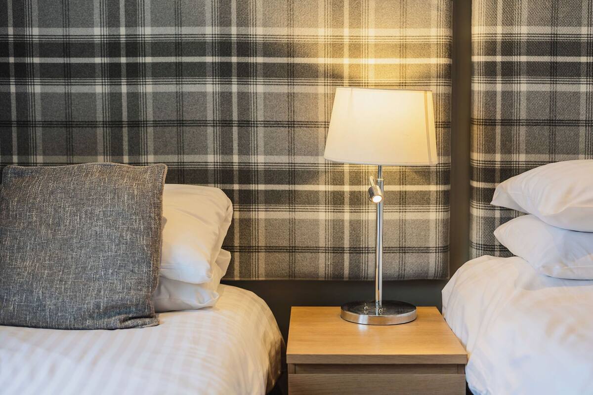 Habitación de hotel con un cabecero estampado en pata de gallo, una lámpara de noche y una cama bien tendida con almohadas.