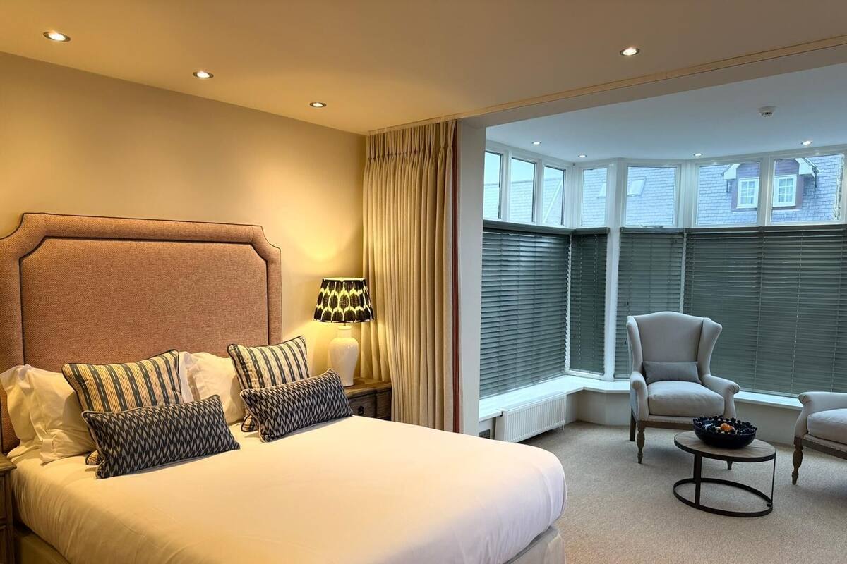 Habitación de hotel con una cama grande, una mesita de noche con lámpara, una silla y vistas al exterior a través de amplias ventanas.