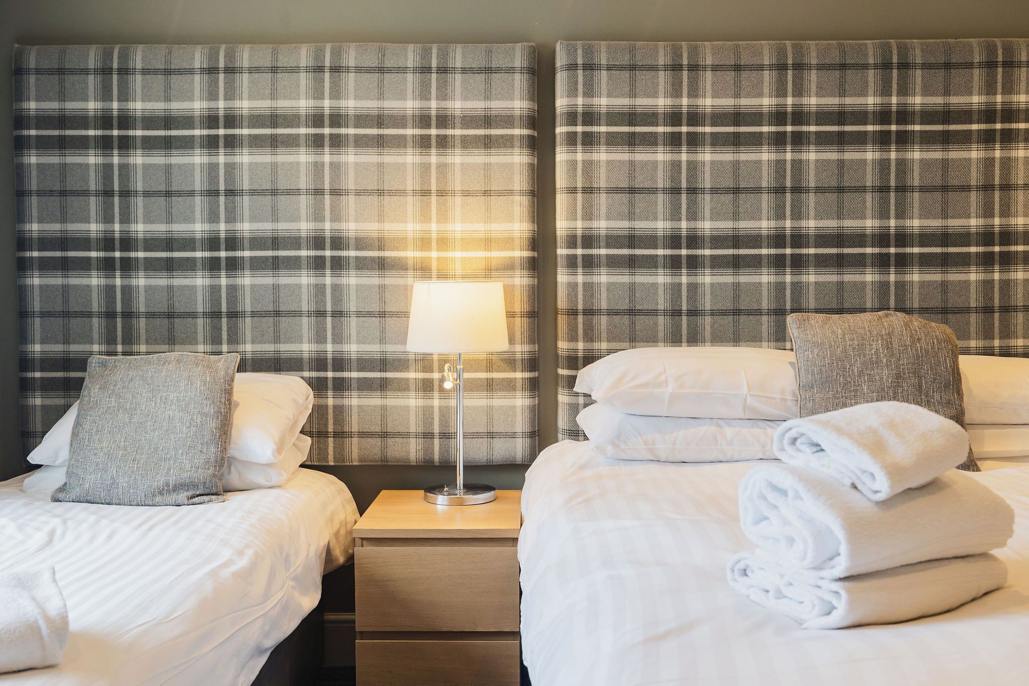 A hotel room with two beds, each with white linens and pillows, a nightstand with a lamp, and plaid-patterned headboards.