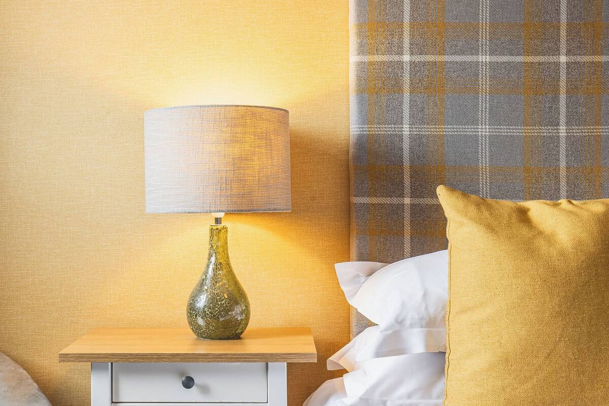 Una cama bien hecha con sábanas blancas, un almohadón amarillo y una lámpara sobre una mesita de noche.