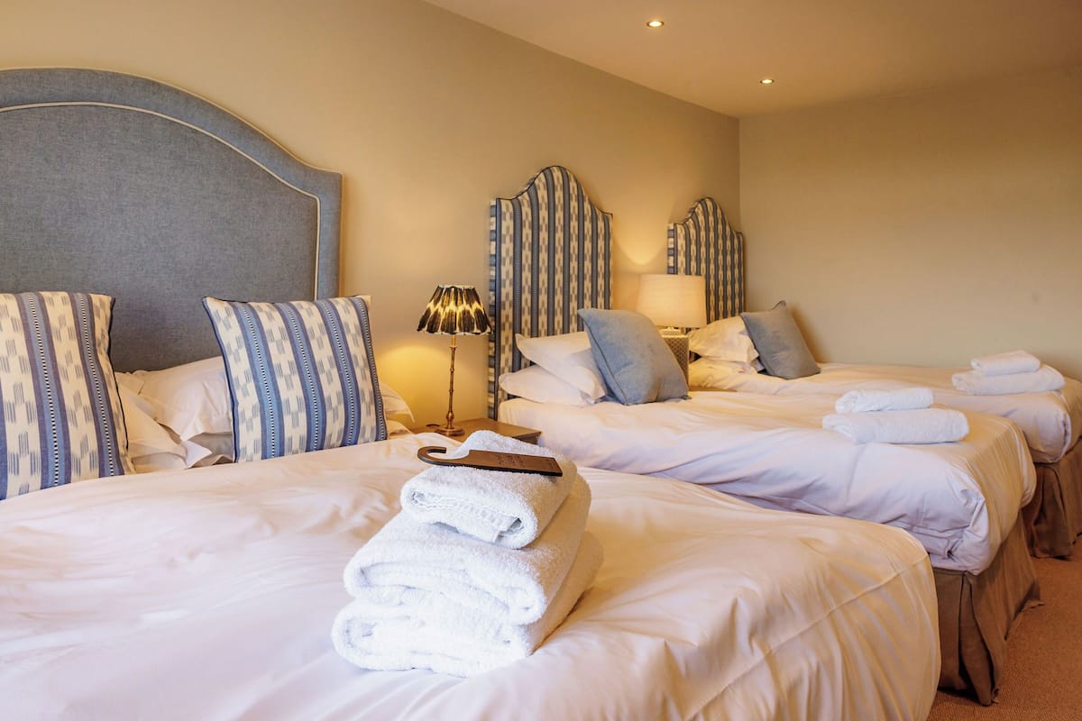 Habitación de hotel con tres camas, cada una con ropa blanca y almohadas a rayas. Se ven toallas dobladas ordenadamente sobre las camas.