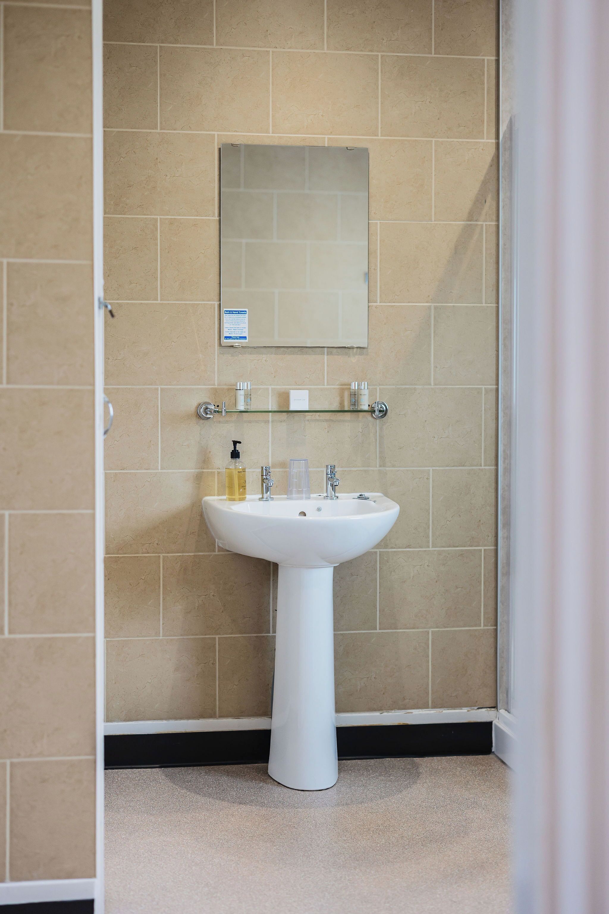 A bathroom with beige tiles, a white sink, a mirror, and a glass shelf with toiletries.