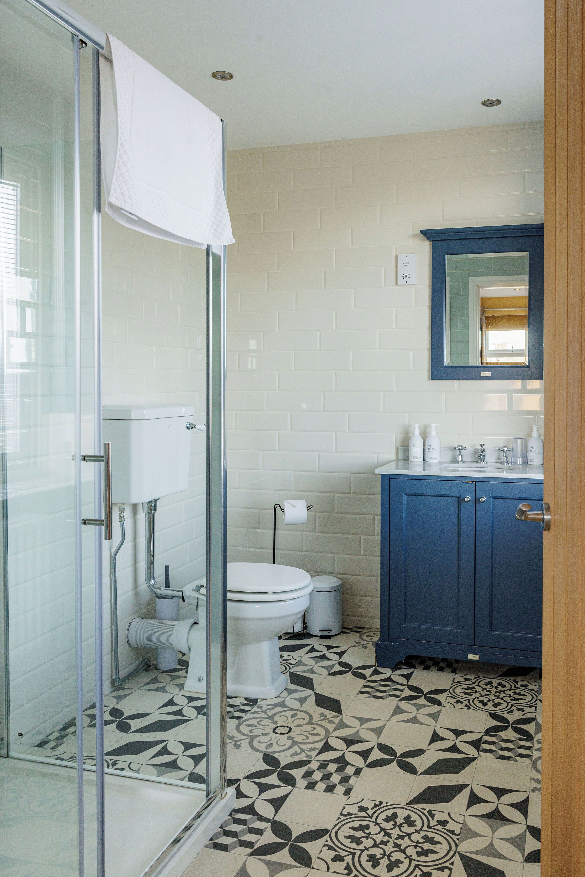 Un baño con cabina de ducha de vidrio, inodoro, lavamanos con mueble azul y baldosas de piso estampadas.