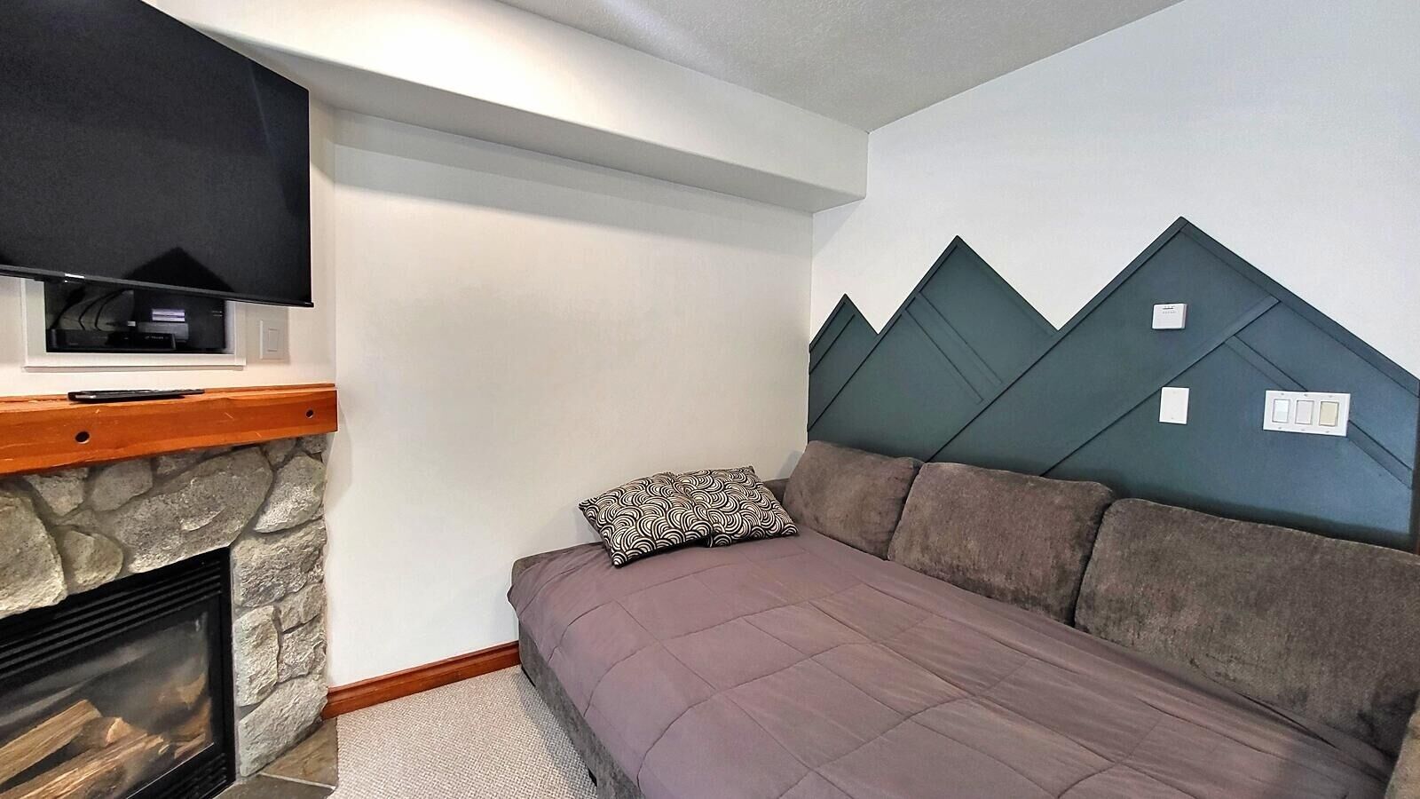 Ein gemütliches Zimmer mit Kamin aus Stein, einem flachbildfernseher darüber, einem Bett mit einem gemusterten Kopfkissen und einer Wand mit geometrischem Dekor.