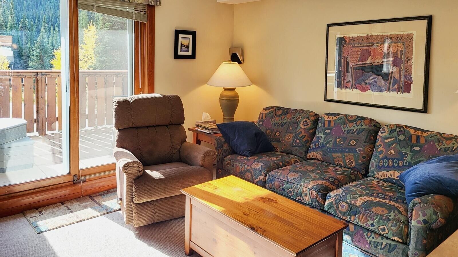 A living room with a brown armchair, a patterned sofa, a wooden coffee table, a lamp, and a framed picture on the wall.