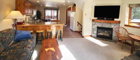 A cozy living room with a sofa, a coffee table, and a fireplace. There is a kitchen area with wooden cabinets and a dining table.