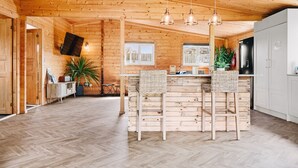 Ein Holzinterieur mit Fischgrätparkett, einer Bar mit Hockern, einem an der Wand befestigten Fernseher und einem Kühlschrank.