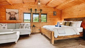 A wooden cabin interior with a bed, sofa, and two windows with views.
