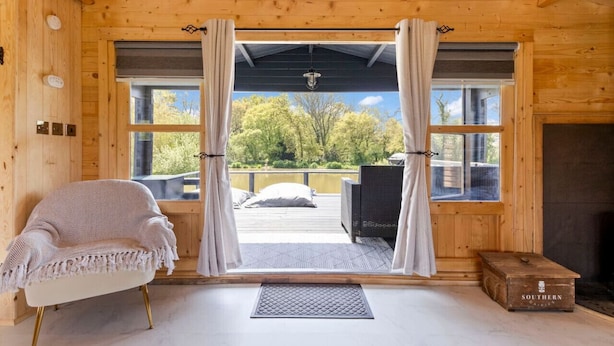 A wooden cabin interior with a cozy seating area, a fireplace, and a view of a deck and trees outside.