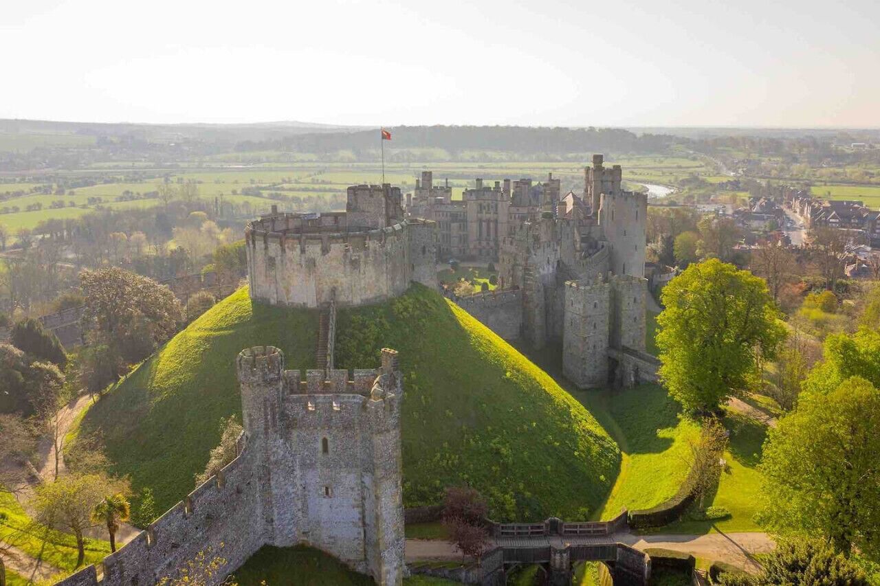 A historic castle with a prominent central tower, surrounded by lush green hills and a small village in the background.