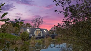 Een klein huis met een schuin dak en een groot raam, omgeven door water en begroeiing.
