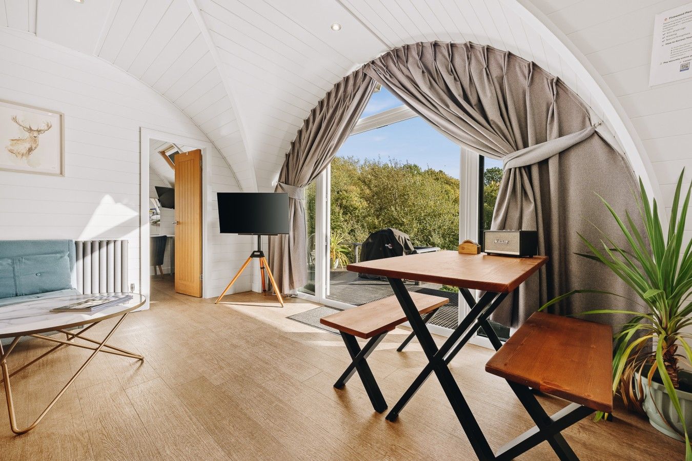 Ein gemütliches Zimmer mit Holzboden, einer Couch, einem Tisch mit Stühlen, einem Fernseher auf einem Stativ und Blick auf Grünfläche.