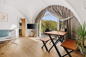Une pièce chaleureuse avec un plancher en bois, un canapé, une table avec des chaises, un téléviseur posé sur un trépied et une vue sur la verdure à l’extérieur.