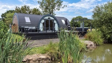 Situated by a calm water body, this house features a curved roof and a prominent front entrance with a glass door and small balcony, surrounded by lush greenery and a well-maintained garden.