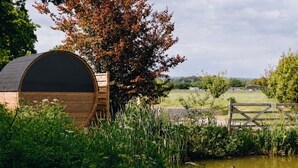 Een houten constructie in landelijke stijl met een grote ronde opening, omgeven door weelderig groen en een vijver met eenden.
