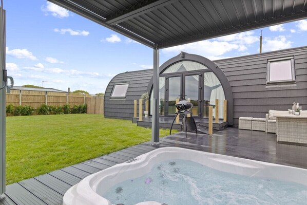 A modern outdoor patio area with a hot tub, a covered seating area, and a large arched window. - Nestledstays - The Barn House (Arundel)