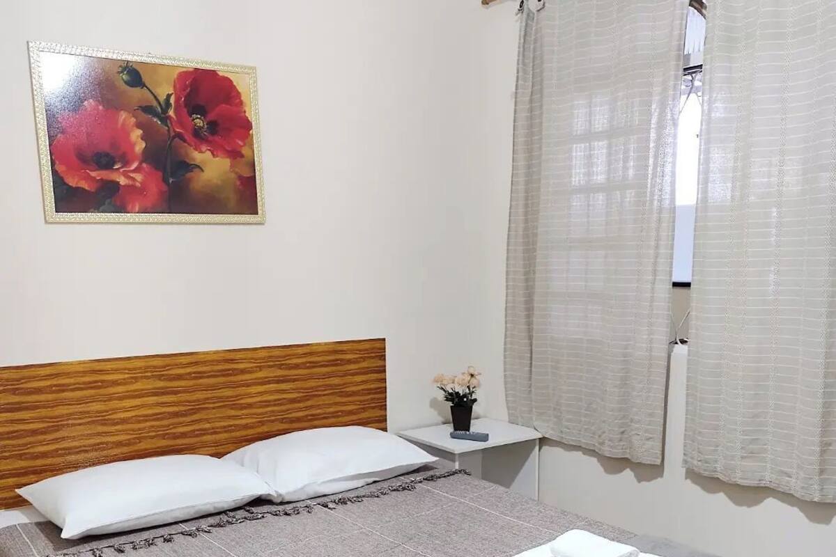 A bedroom with a wooden headboard, a bed with two pillows, a bedside table with a vase of flowers, and a window with sheer curtains.