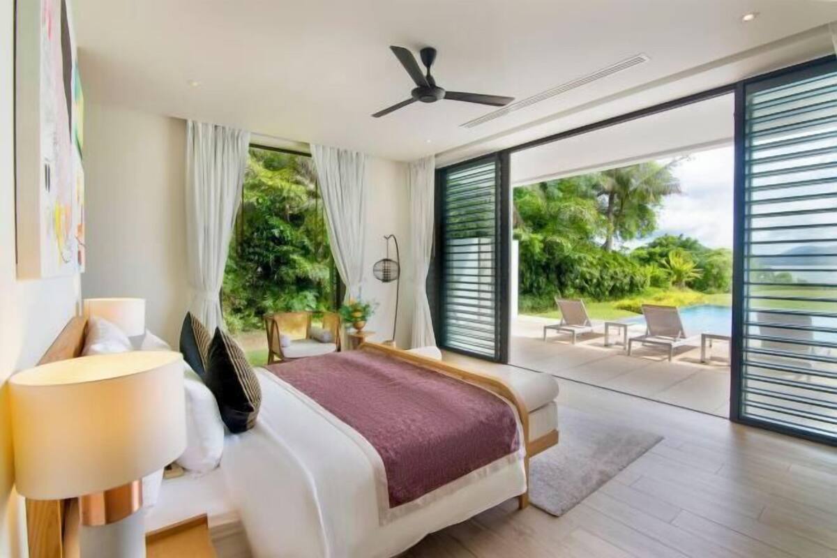 Une chambre avec un lit, un ventilateur de plafond, une table de chevet, une lampe et une vue sur un espace extérieur comprenant une piscine et des chaises.