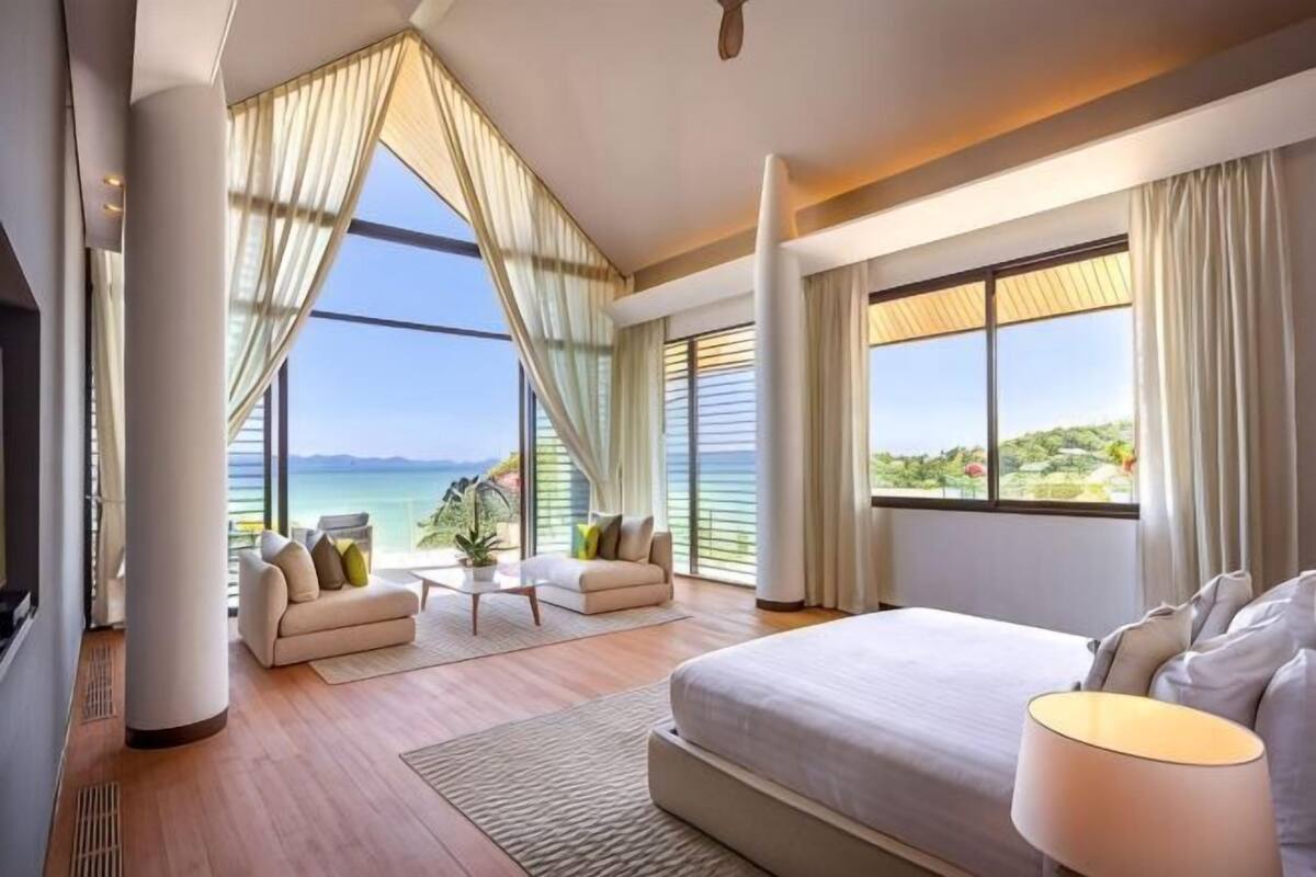 Une chambre d’hôtel moderne, dotée d’un grand lit, d’un canapé, d’une table basse et d’un téléviseur. La chambre dispose de grandes fenêtres offrant une vue sur la mer.