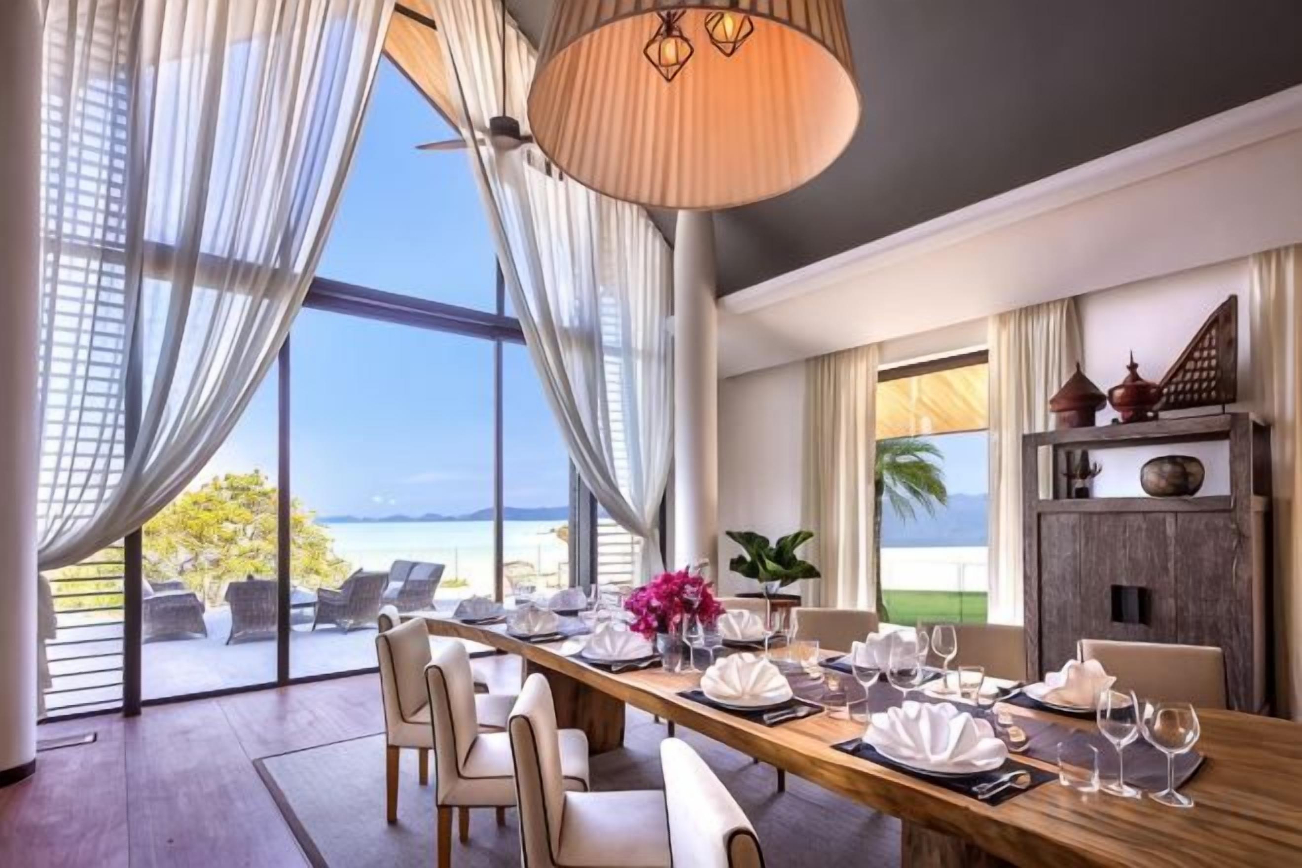 A dining area with a long table set for a meal, large windows with curtains, and a view of the outdoors.