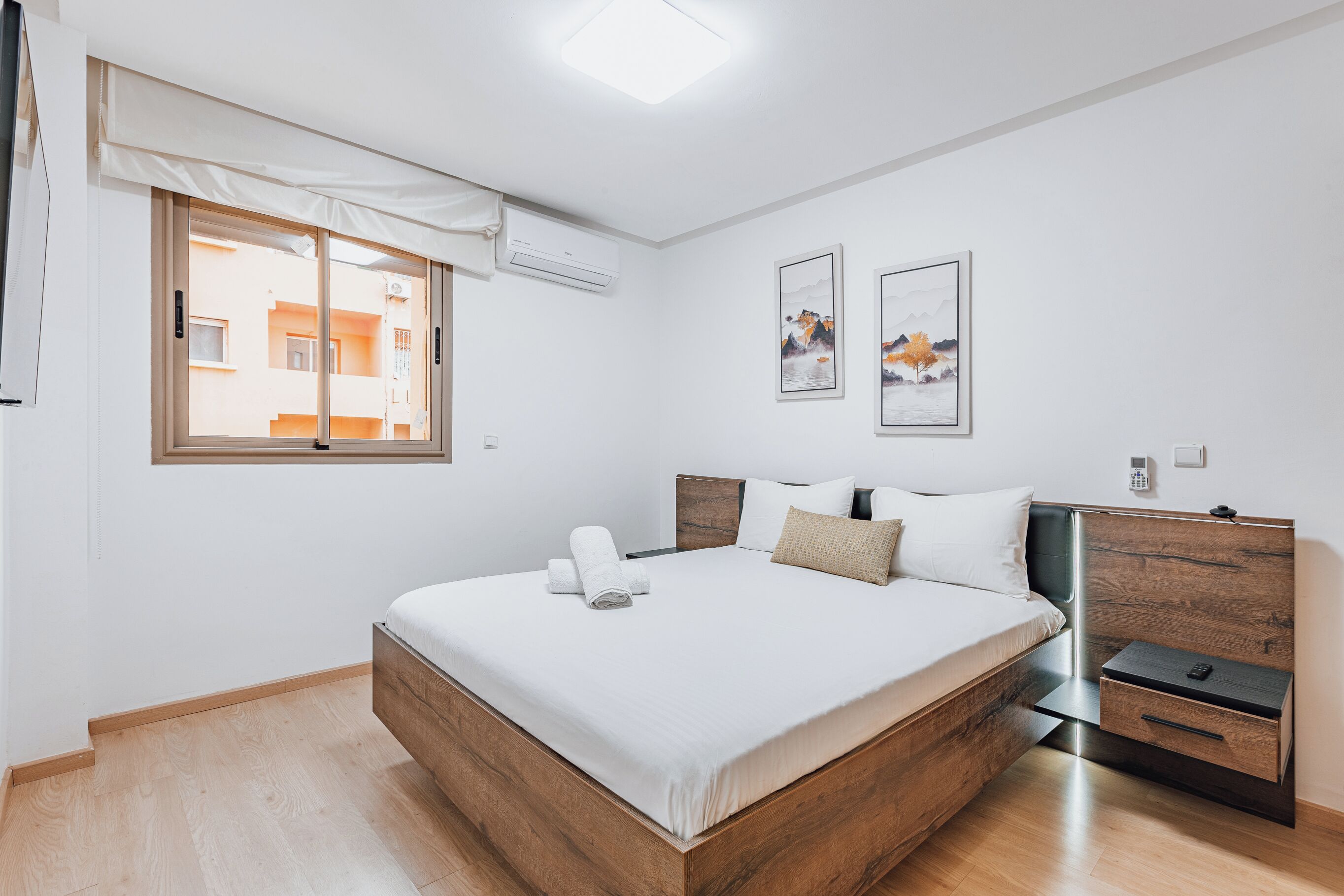 A modern bedroom with a wooden bed, a bedside table, and a window with a view of buildings.