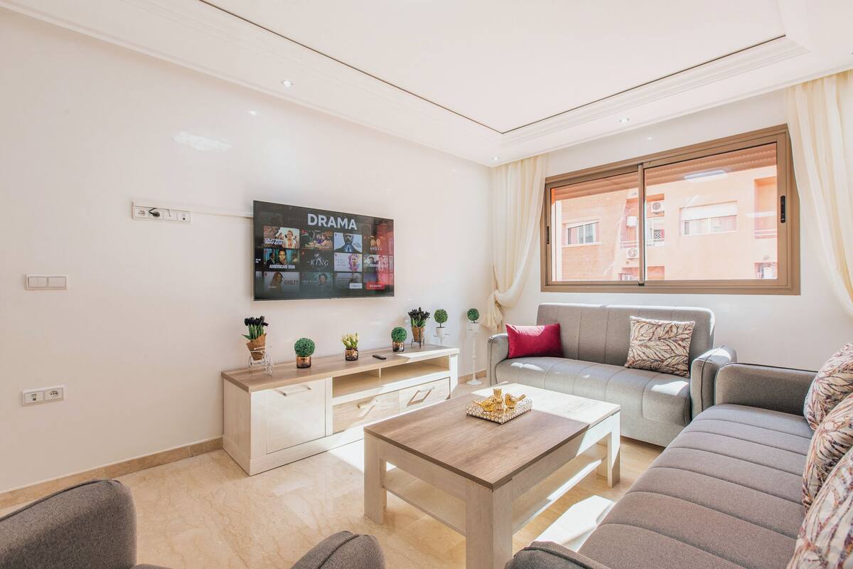 Un salon moderne avec un canapé gris, une table basse en bois et un téléviseur fixé au mur, diffusant une chaîne consacrée aux séries dramatiques.