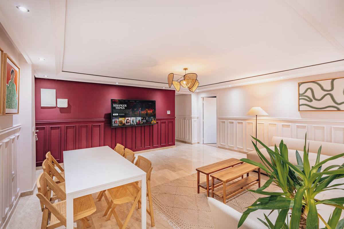 Un salon spacieux avec un canapé blanc, une table à manger en bois et des chaises. Un téléviseur est fixé au mur, et une plante se trouve dans un coin.