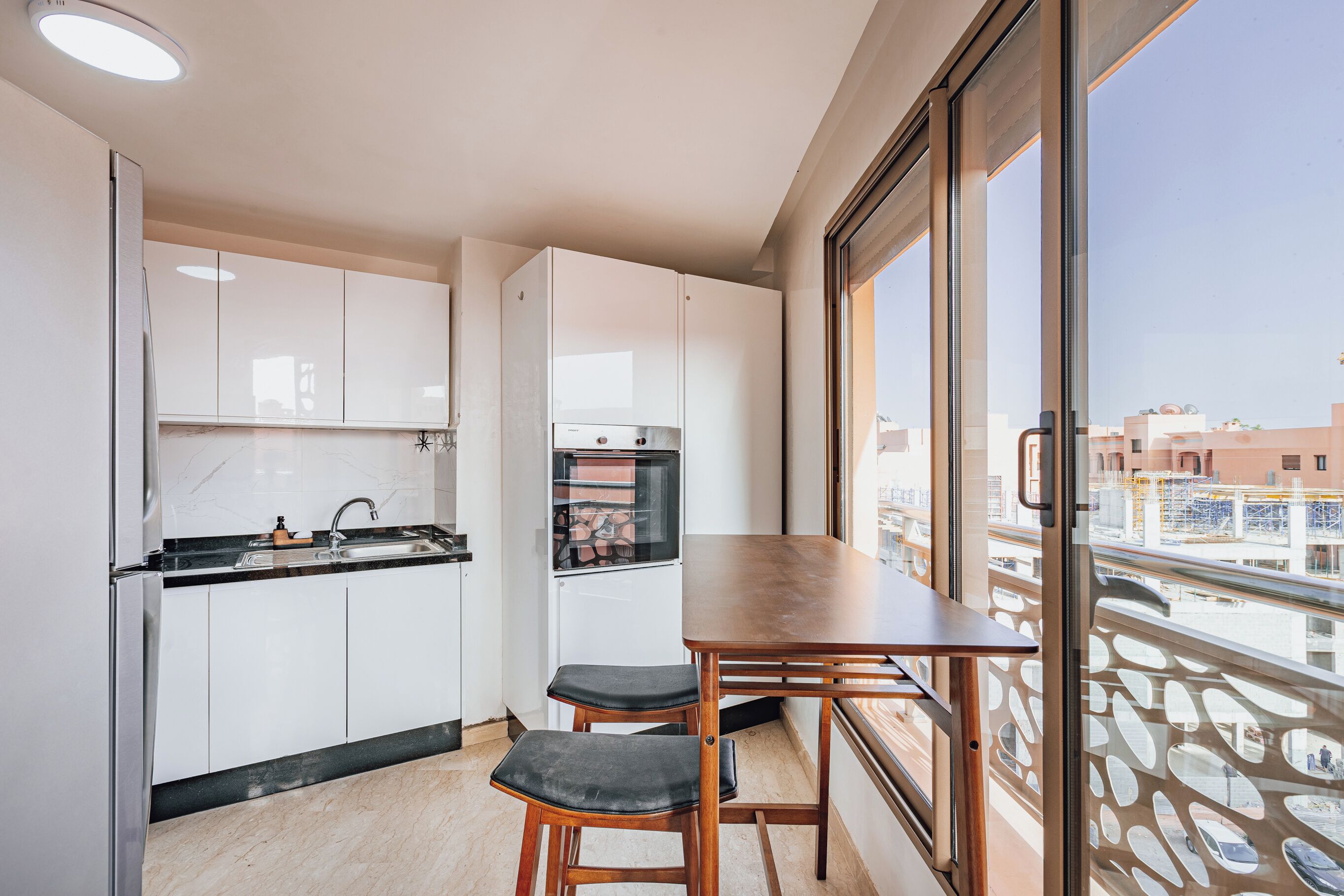 A compact kitchen with white cabinets, a black countertop, and a wooden table with two stools. A refrigerator and oven are visible. The room has large windows offering a view of buildings outside.