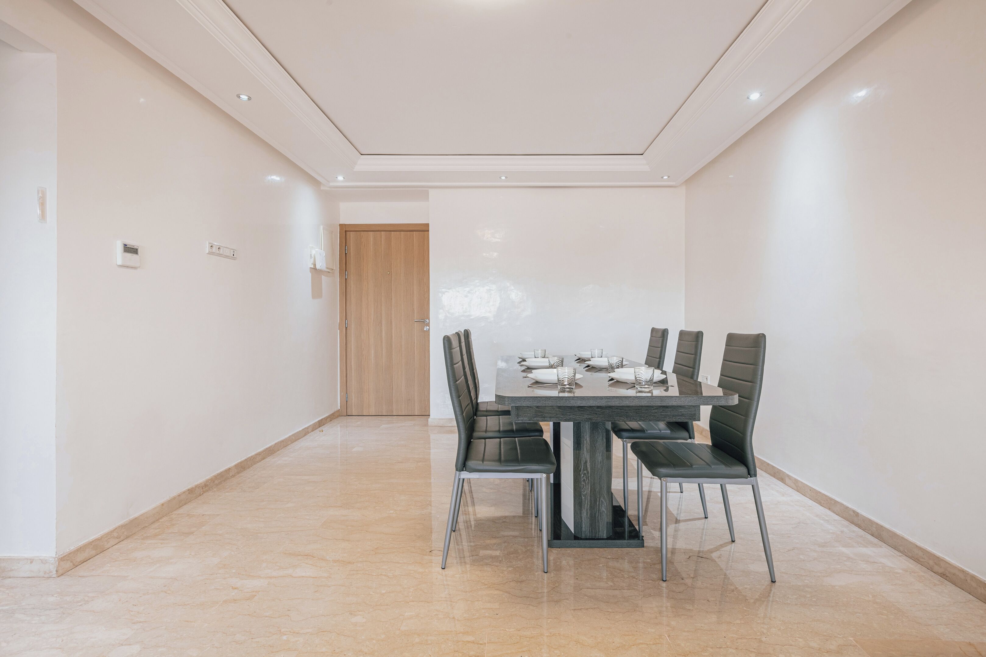 A modern dining area with a rectangular table and six chairs, a wooden door in the background, and a tiled floor.