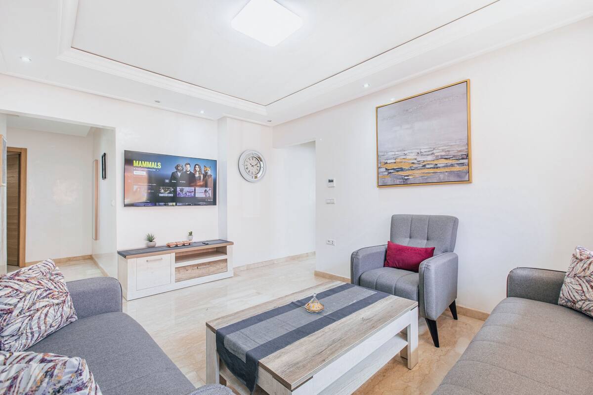 Un salon moderne avec des canapés gris, une table basse, un téléviseur diffusant une émission sur les « mammifères », une horloge et un tableau accroché au mur.