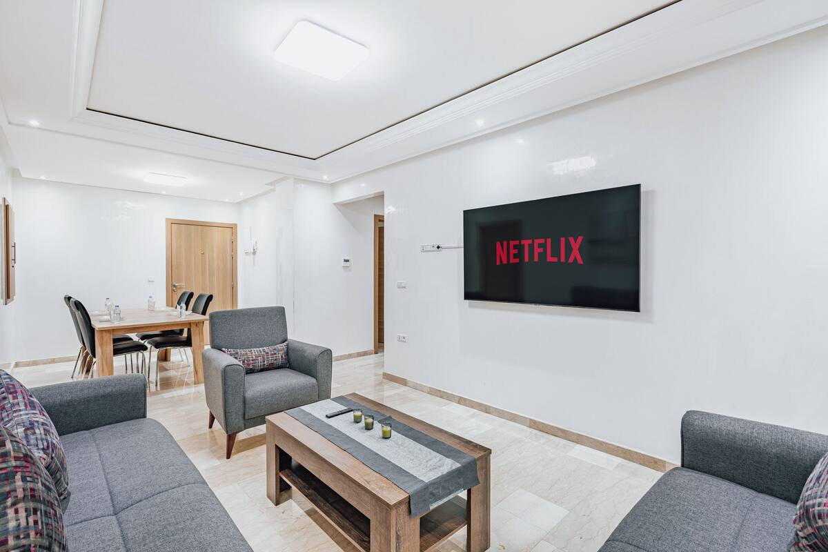 Un salon moderne avec une table basse en bois, des canapés gris, un coin repas avec une table et des chaises en bois, et un téléviseur fixé au mur qui affiche le logo de Netflix.